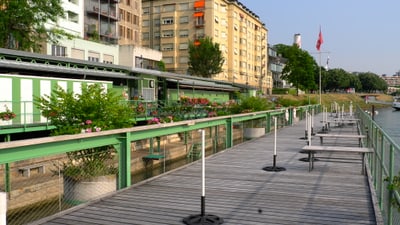 Holzsteg mit Sonnenschirmhalterungen und Blumentöpfen, dahinter die Garderobengebäude und Wohnhäuser am Rheinufer.