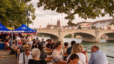 Menschen sitzen an Tischen am Flussufer mit Brücke im Hintergrund.