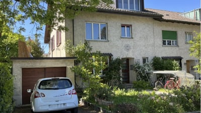 Zwei beige Häuser mit Garagen und einem Auto im Hof bei sonnigem Wetter.