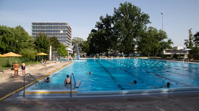 Aussenpool mit Schwimmern und Gebäuden im Hintergrund.