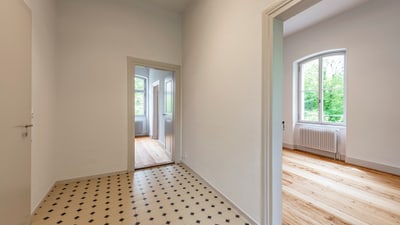 Leerer Flur mit Fliesenboden und Blick in zwei Räume mit Holzböden und Fenstern.