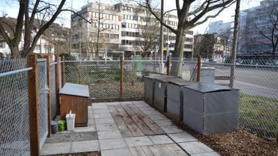 Eingezäunter Bereich mit Behältern und Gebäuden im Hintergrund.