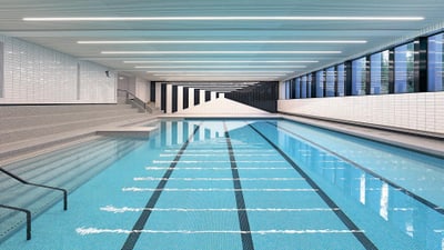 Schwimmbecken mit einem ganzseitigen Treppeneinstieg. Schwimmhalle mit weissen Plättli und heller Beleuchtung an der Decke.