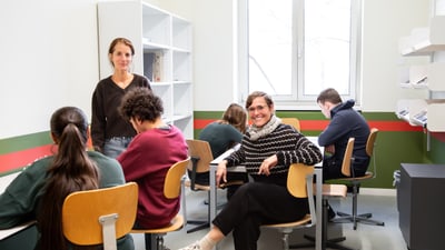 Menschen arbeiten in einem Klassenzimmer an Tischen, lächelnde Person in der Mitte.