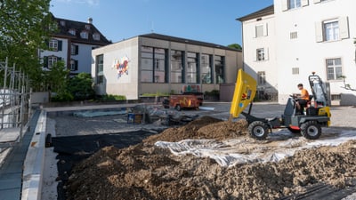 Bauarbeiter bedient Bagger auf Baustelle vor Gebäuden.