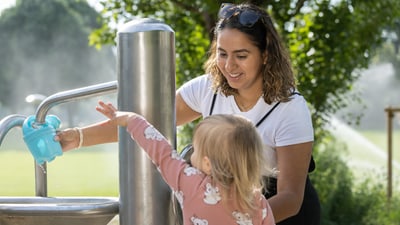 Frau hilft Kind an einem Wasserspender im Park.