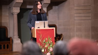 Frau hält Vortrag an einem Rednerpult vor Publikum.