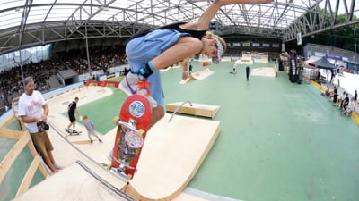 Skateboarder beim Trick in einer Skatehalle mit Publikum.