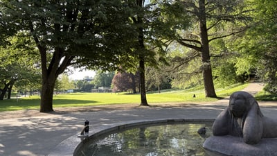 Skulptur an einem Brunnen in einem Park mit Bäumen und Rasenfläche.