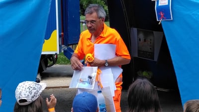 Mann in orangefarbener Kleidung spricht zu Kindern unter Zeltplane.",