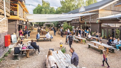 Menschen sitzen und stehen in einem Innenhof mit Holzbauten und Bänken.