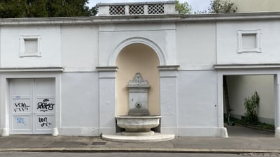 Weisse Wand mit Brunnen und Graffiti, offene Durchgänge seitlich.