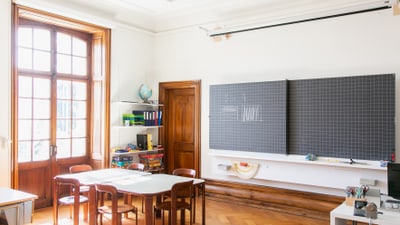 Hell beleuchtetes Klassenzimmer mit Tafel und Holztischen.