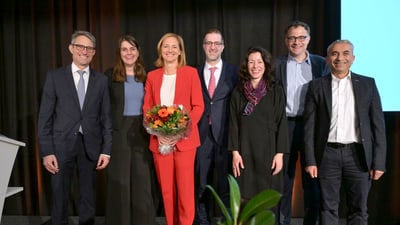 Gruppe von sieben Personen, eine hält Blumenstrauss, lächeln auf Bühne.",