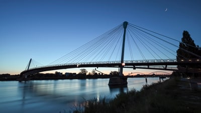 Hängebrücke über ruhigen Fluss bei Dämmerung mit Mondsichel.