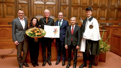 Sechs Personen in formeller Kleidung mit Blumenstrauss und Urkunde in Holzzaal.