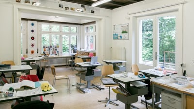 Klassenzimmer mit Tischen und Stühlen, helle Fenster.