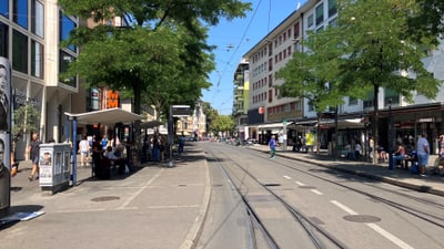 Stadtstrasse mit Gehwegen, Bäumen und Geschäften bei sonnigem Wetter.