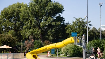 Gelbe Wasserrutsche in einem Freibad mit Bäumen im Hintergrund.