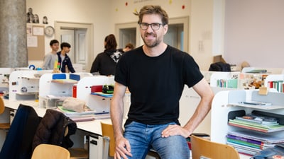 Heiko Vollmer sitzt lächelnd in einem Klassenraum, andere Personen im Hintergrund.