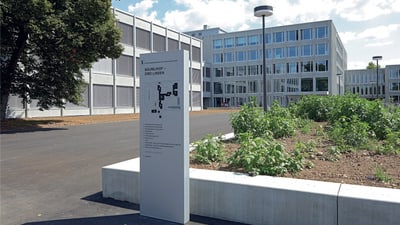 Schulhaus, davor ein asphaltierter breiter Weg und eine Tafel auf der Steht "Mäumlihof - Drei Linden" und ein Gebäudeplan der Schule. 