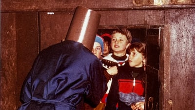 Mensch mit Eimerkopf spricht zu Kindern an der Tür.