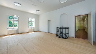 Leerer, heller Raum mit Holzfussboden und Fenstern, offene Tür zu Flur.