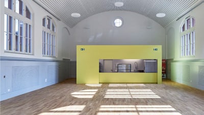 Grosser, leerer Saal mit Holzboden und hohen Fenstern in einem modern renovierten Gebäude.