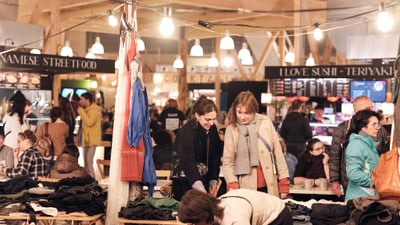 Markt mit Menschen, Verkaufsständen, Essen und Kleidung, beleuchtet von Lichtern.