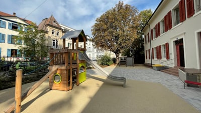 Spielplatz mit Rutsche bei Gebäuden und Baum im Hintergrund.