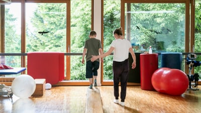 Physiotherapeutin hilft einem Jungen beim Balancieren auf Balken im Therapieraum.