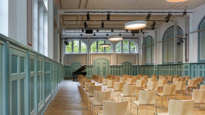 Aula mit Stuhlreihen. Vor der Stuhlreihen am Ende des Raumes steht ein schwarzer Flügel.