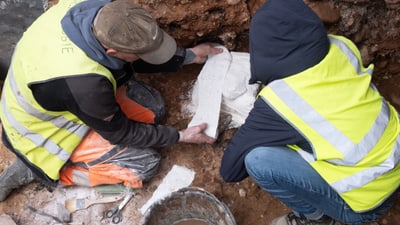 Archäologen legen Fund frei in Grube mit gelben Westen.