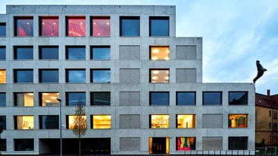 Abgestuftes Gebäude aus Beton, durch dessen Fenster Licht nach aussen dringt und davor eine Laterne und ein schmaler Baum.