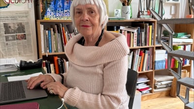 Eine Person sitzt an einem Schreibtisch mit Bücherregalen im Hintergrund.