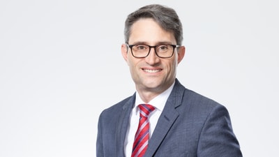 Mann im Anzug mit roter Krawatte und Brille lächelt.