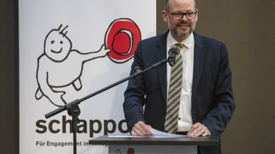 Ein Mann spricht am Pult aauf der Bühne vor einem schappo Plakat.