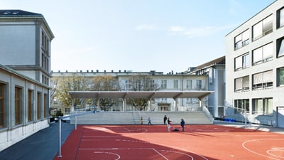 Schulhof mit rotem Basketballplatz und Kindern, umgeben von Schulgebäuden.