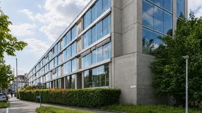 Modernes Glasgebäude an einer Strasse mit Bäumen und Büschen.