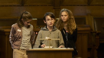 Drei Kinder stehen zusammen in Saal, sprechen in ein Mikrofon.