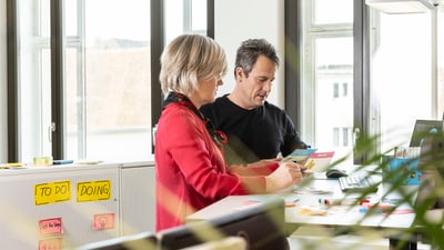 Zwei Personen arbeiten an einem Schreibtisch mit Aufgabenlisten an der Wand.