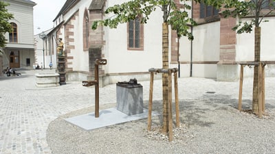 Platz mit zwei Bäumen und Archaeoskop aus Bronze vor einem alten Gebäude.