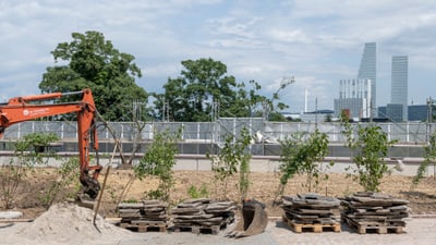 Baustelle mit Bagger und gestapelten Materialien, im Hintergrund grüne Bäume und Hochhäuser.