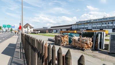 Gartenzaun, Strasse und Lagerbereich mit Holzstapeln vor einem Gebäude.