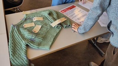 Grüner Pullover mit Etiketten auf Tisch neben stehenden Personen.",