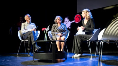 Drei Personen in Diskussion auf Bühne mit Grafik im Hintergrund.