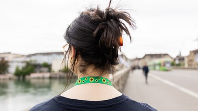 Rückansicht einer Person mit Dutt, die auf einer Promenade geht. Sie trägt einen grünen Bändel mit gelben Sonnenblumen um den Hals.