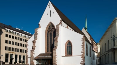 Historische Kirche mit weissen Wänden und hohem spitzen Dach zwischen modernen Gebäuden.