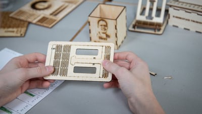 Hände halten ein Holzbauteil auf einem Tisch mit weiteren Bauteilen.