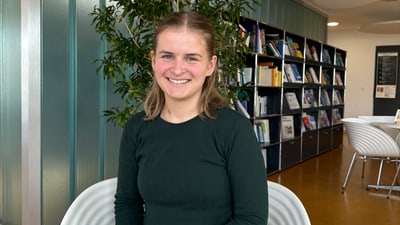 Person sitzt in modernem Raum mit Bücherregal im Hintergrund.